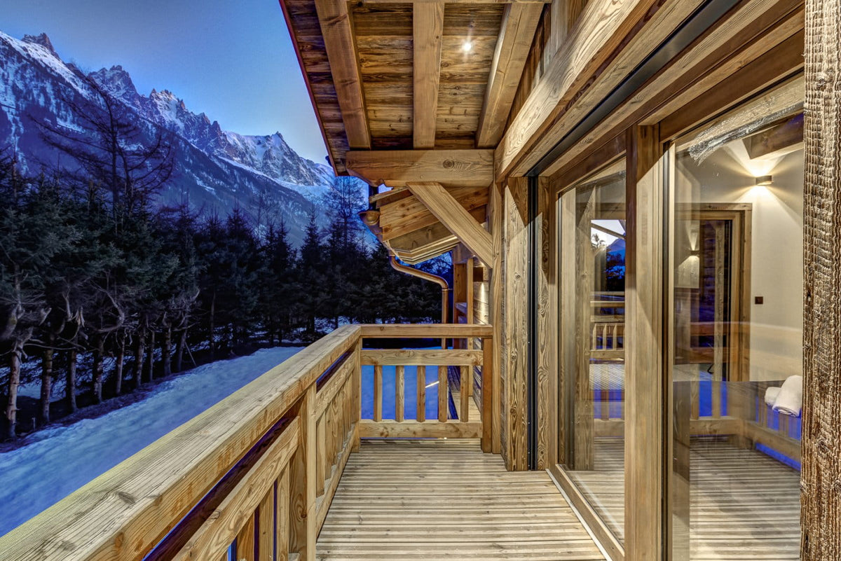 A wooden chalet balcony overlooking a snowy mountain landscape at dusk, with large glass doors, warm interior lighting, and snow-covered peaks in the background.