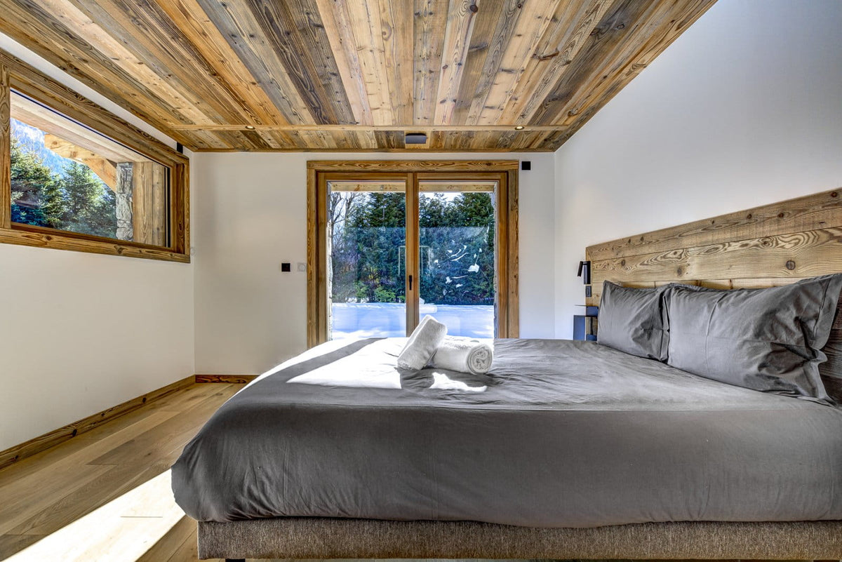 A modern bedroom featuring a large bed with gray bedding, a rustic wooden headboard and ceiling, white walls, and a large window looking out onto a snowy outdoor scene.