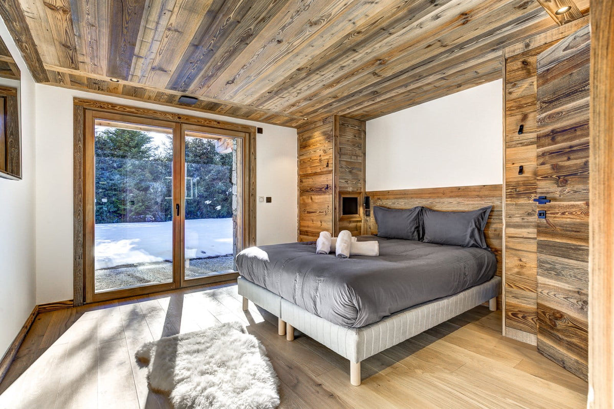 A modern bedroom with a bed dressed in gray bedding, set against rustic wood-paneled walls and a matching wood ceiling. Large glass doors on the left reveal a snowy outdoor view.