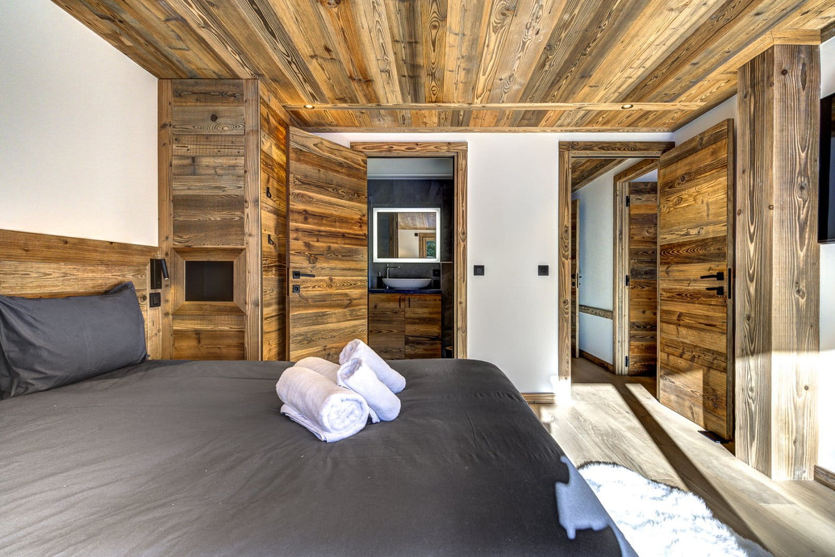 A modern bedroom interior with a bed dressed in dark gray bedding, white towels on top, rustic wood-paneled walls and ceiling, and open doors revealing a bathroom and hallway.