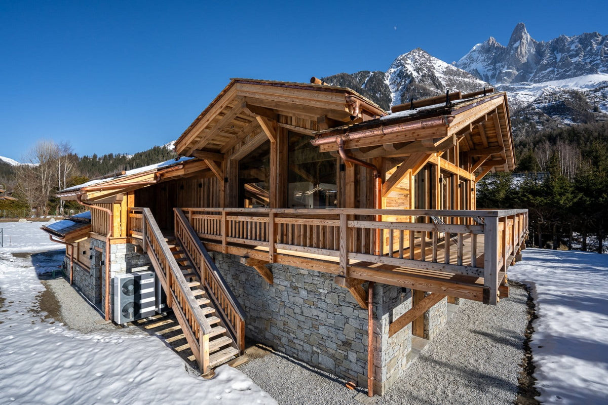 A rustic wooden chalet-style mountain house with stone foundations, large windows, and a wooden deck, set in a snowy landscape with snow-capped peaks under a clear blue sky.