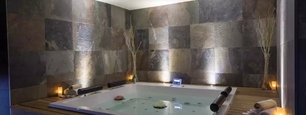 An indoor spa-style hot tub or bathtub set into a stone-tiled room with warm ambient lighting, candles, and decorative branches.