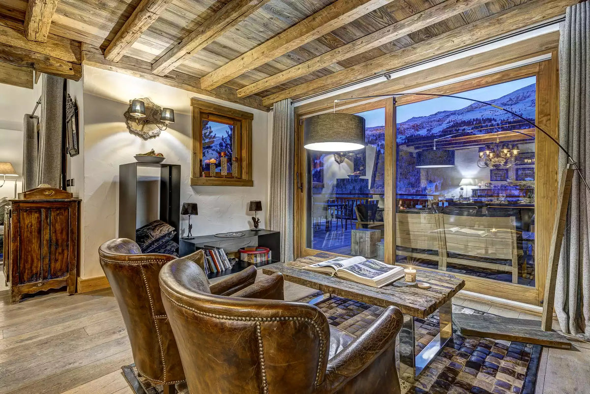 A luxury mountain chalet living room featuring rustic wooden ceiling beams, two brown leather armchairs, a wooden dining table, and a large floor-to-ceiling window showcasing a snowy mountain landscape at dusk.
