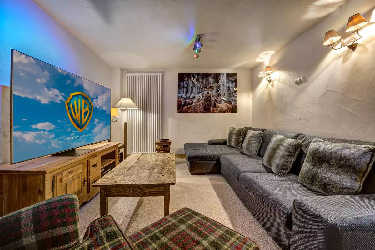A modern living room setup featuring a large flat-screen TV displaying the Warner Bros. logo, a gray sectional sofa with textured pillows, a wooden coffee table, and plaid accent chairs.