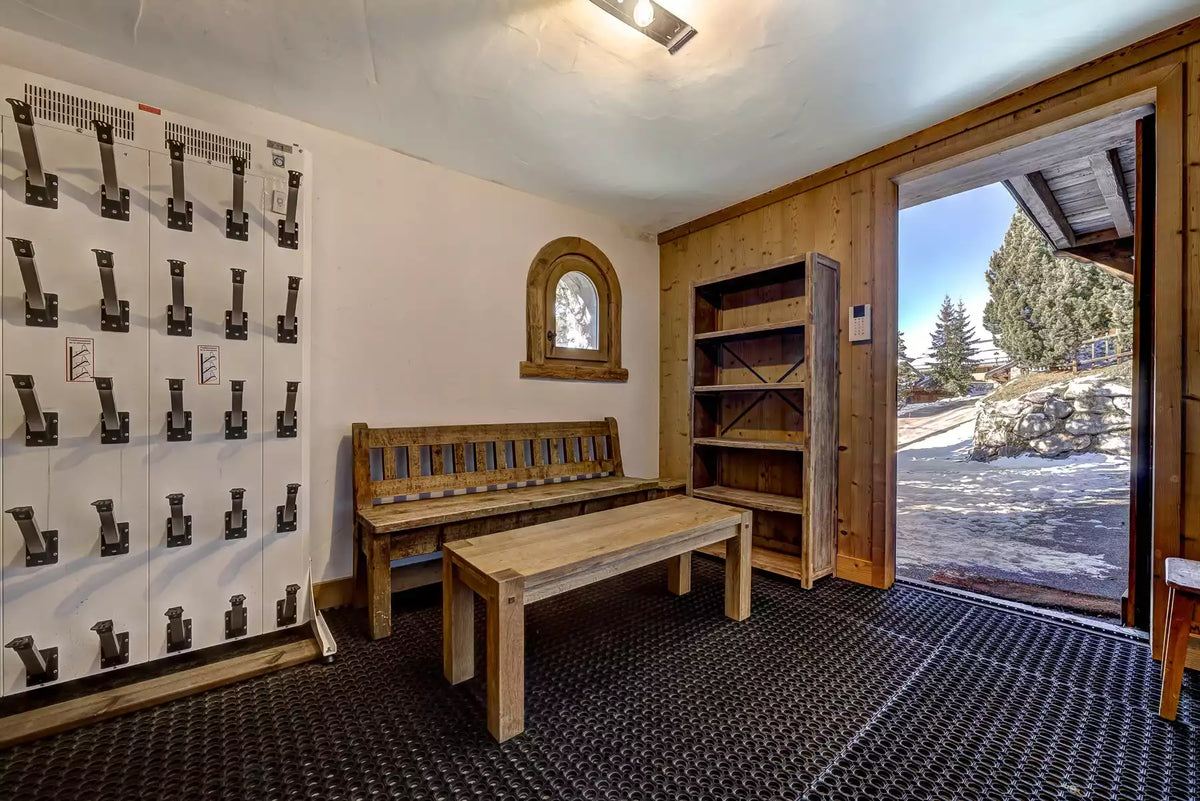 A rustic wooden bench and matching table in a lodge-style interior with a large open door showing a snowy outdoor mountain view. A white wall on the left features a grid of black ski boot lockers, and a wooden bookshelf stands against the back wall.