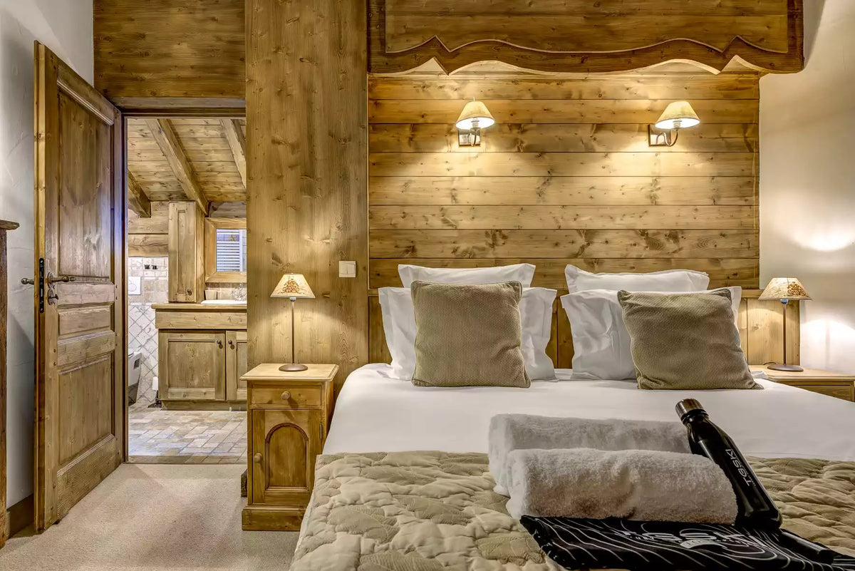 A rustic bedroom interior with a large bed dressed in white linens and beige pillows, set against a wood-paneled wall. A doorway on the left reveals a bathroom, and two bedside tables with lamps flank the bed.