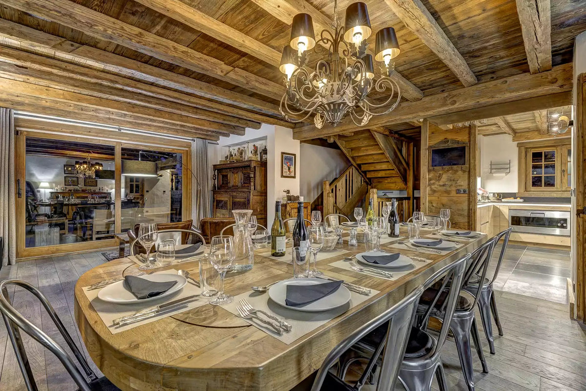 A rustic wooden dining table set for six with plates, wine glasses, and cutlery, surrounded by metal chairs in a chalet-style dining room with exposed wood ceiling beams and a chandelier.