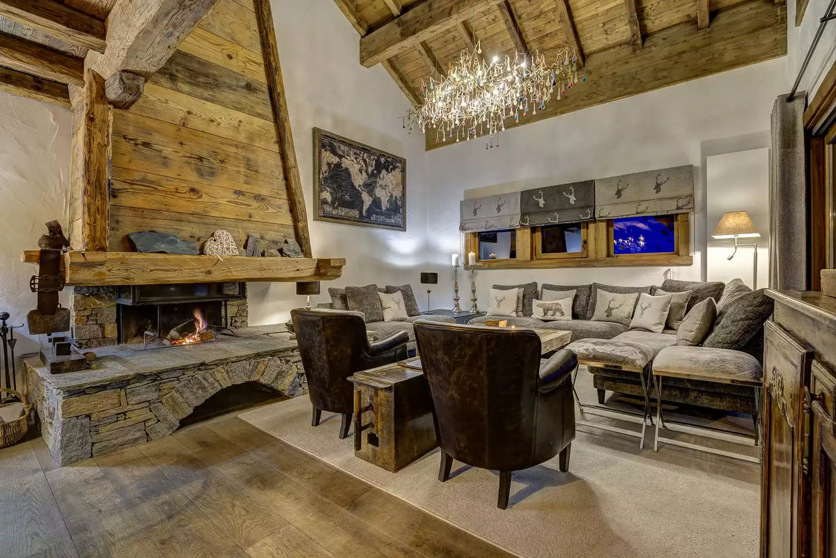 A luxurious rustic chalet living room featuring a large stone fireplace with a wood-burning stove, a plush sectional sofa, two dark leather armchairs, a crystal chandelier, and exposed wooden ceiling beams.