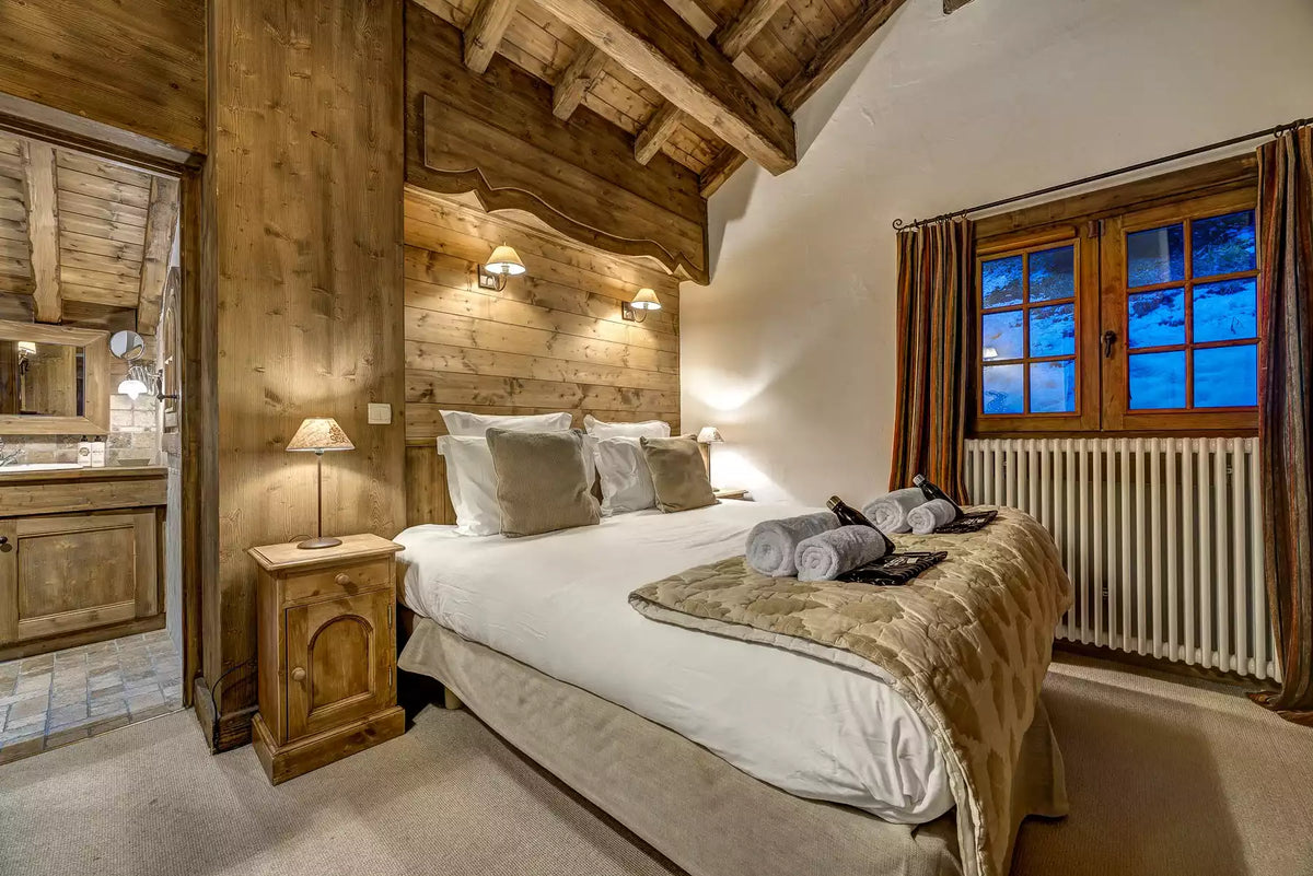 A cozy alpine-style bedroom featuring a large bed with white linens, a rustic wood-paneled headboard, matching wooden nightstands, warm wall sconces, and a window with brown curtains showing a blue evening view outside.