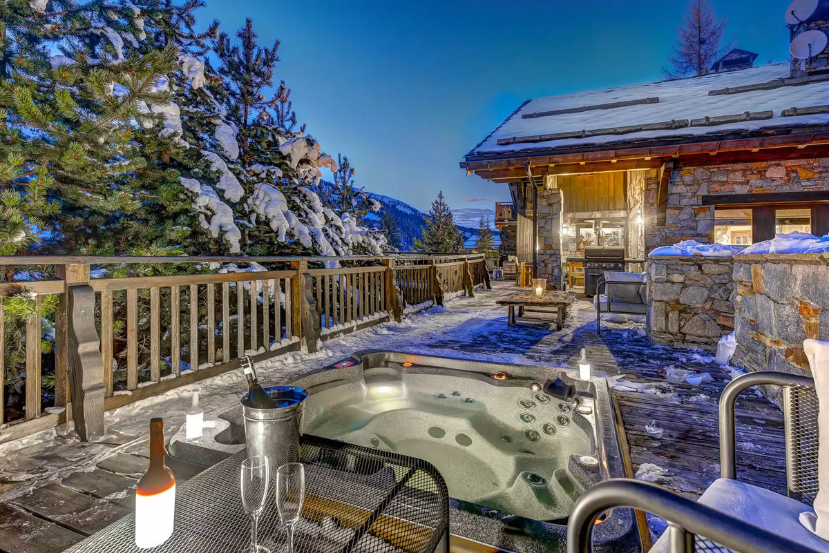 An outdoor hot tub sits on a snowy patio beside a rustic stone chalet at dusk, with snow-covered trees and mountains in the background. A bottle, ice bucket, and two wine glasses are visible on a table in the foreground.