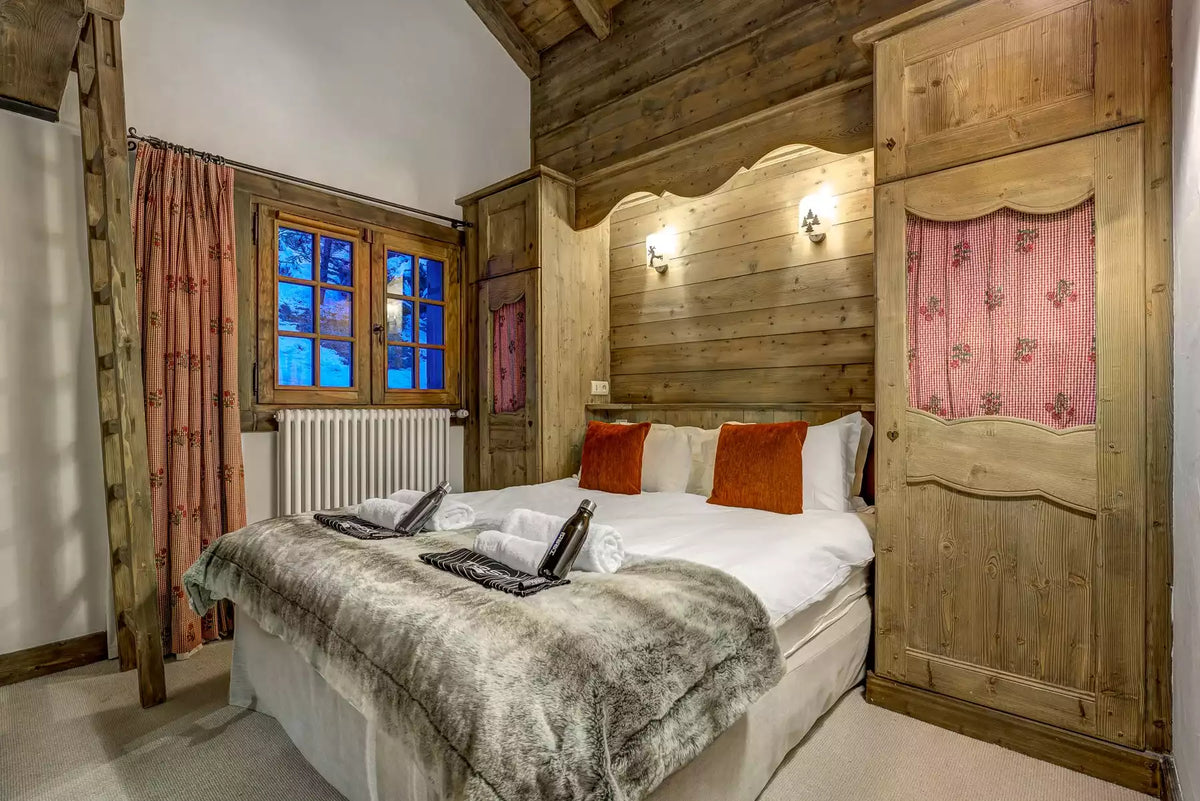 A rustic alpine-style bedroom featuring a large wooden wardrobe, a wooden headboard, a bed with white bedding and orange accent pillows, and pink patterned curtains by a window.