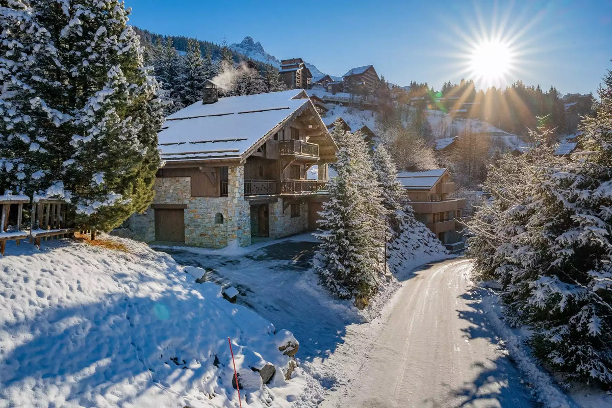 A snowy mountain chalet with stone and wood exterior walls, surrounded by snow-covered evergreen trees and a sunlit snow path under a bright blue sky.