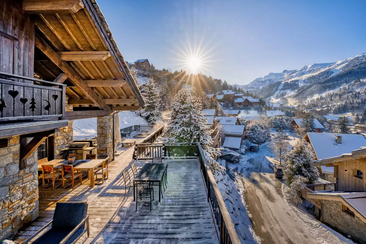 A luxurious alpine chalet with a large wooden deck overlooking a snow-covered mountain village under a bright sun in a clear blue sky.