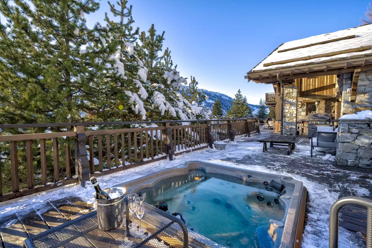 A luxury outdoor hot tub on a snowy mountain chalet patio, featuring a champagne bucket and glasses, with snow-covered pine trees and a wooden cabin in the background.