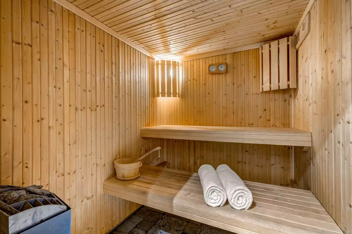 A wooden sauna interior with light natural wood paneled walls and ceiling, a built-in wooden bench, a hanging wooden bucket, two rolled white towels on the bench, and a steam generator on the floor.