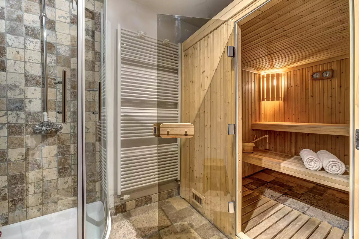 A modern bathroom featuring a glass shower enclosure with stone tile walls and a wooden sauna room with built-in benches, warm lighting, and rolled towels.