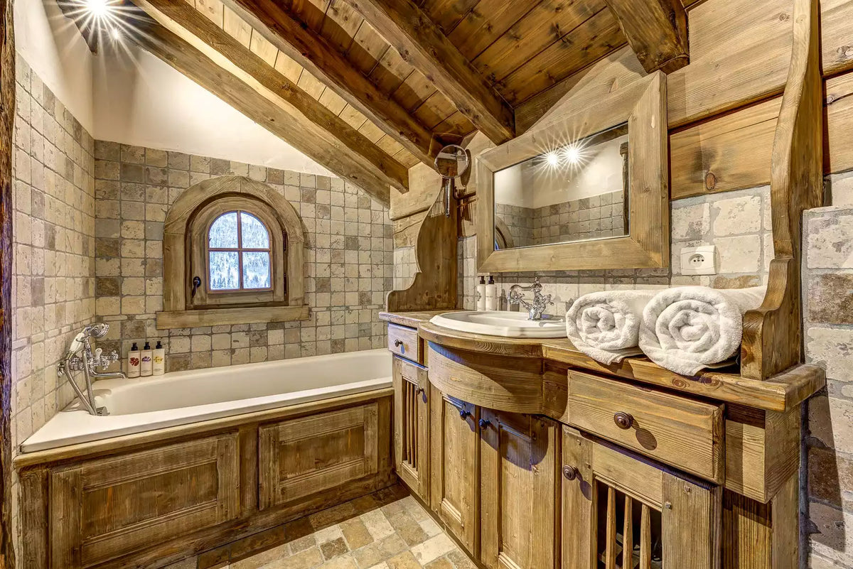 A rustic cabin-style bathroom featuring a large wooden vanity with an integrated sink, a white bathtub, stone tile walls, a small arched window, and rolled white towels on the counter.