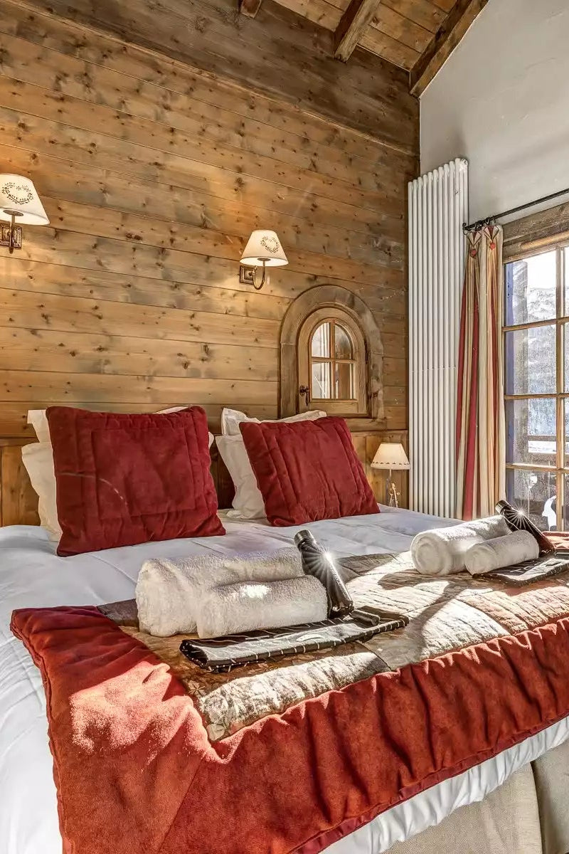 A cozy mountain chalet bedroom featuring a double bed with white linens and red pillows, set against a rustic wood-paneled wall. A wooden ladder leans against a white wall next to a window with red-and-white striped curtains.