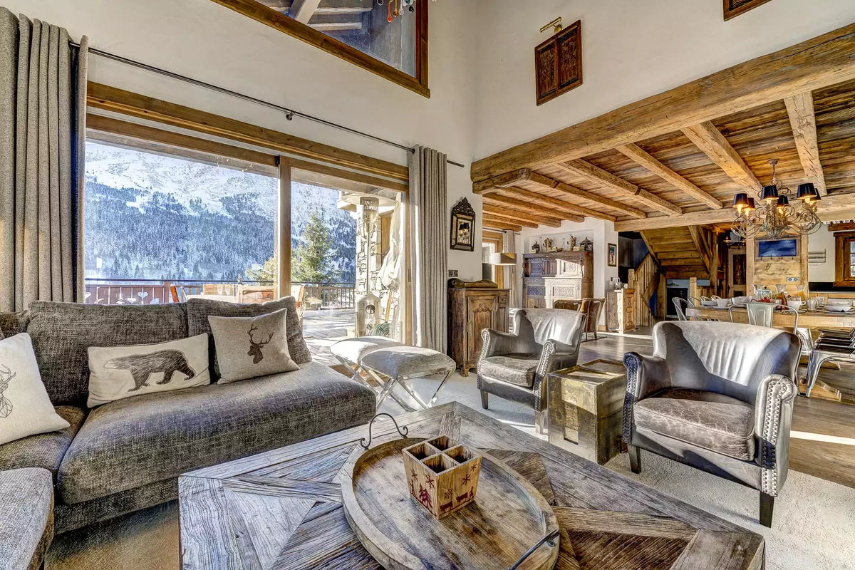 A luxury mountain chalet living room featuring a gray fabric sofa with decorative pillows, a rustic wooden coffee table, leather armchairs, and large windows showcasing a scenic mountain view.
