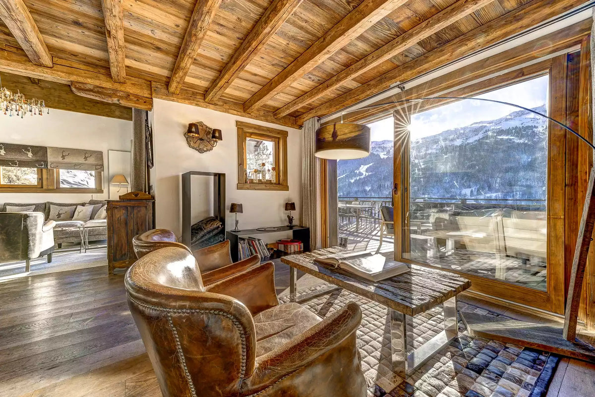 A rustic alpine chalet interior with exposed wooden ceiling beams, large floor-to-ceiling windows, leather armchairs, a wooden writing desk, and a mountain view outside.