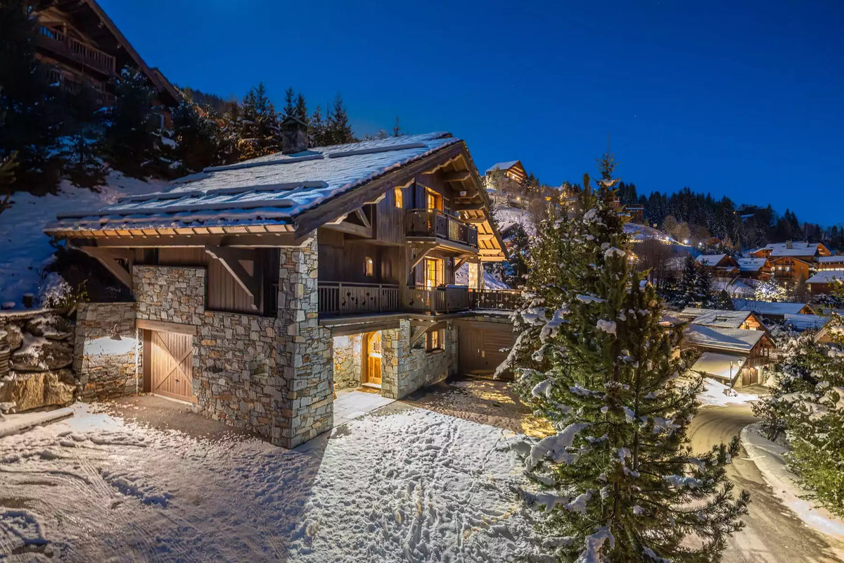 A luxury chalet-style mountain villa with stone and wood exterior walls, snow-covered roof and driveway, warmly lit windows, surrounded by snow-laden pine trees in a snowy alpine village at dusk.