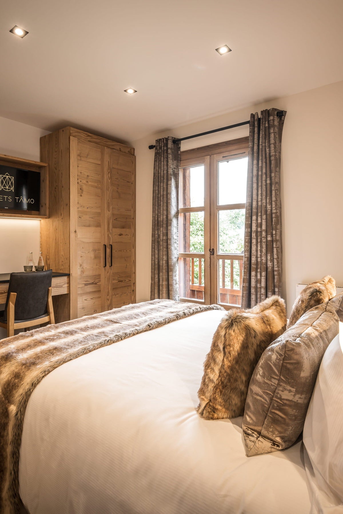 A rustic bedroom interior with a large bed dressed in white linens and patterned pillows, wooden wardrobes, a desk nook, and a window with curtains.