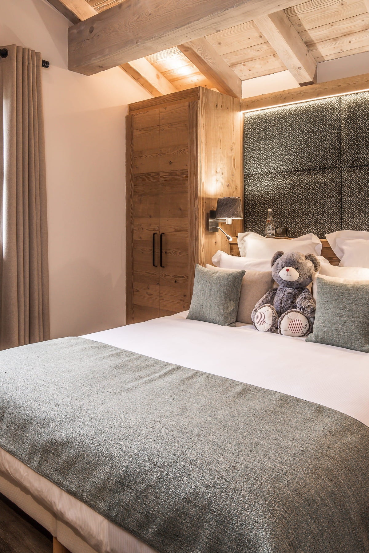 A cozy bedroom interior featuring a neatly made bed with white linens, gray-green throw and pillows, a plush toy, a dark textured headboard, wooden ceiling beams, a window with curtains, and a large wooden wardrobe.