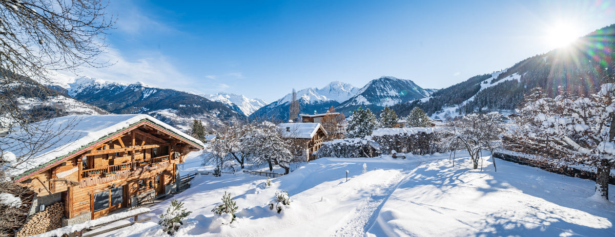 A snowy mountain village scene with wooden chalets, snow-covered trees, distant snow-capped peaks, and a bright sun in a clear blue sky.