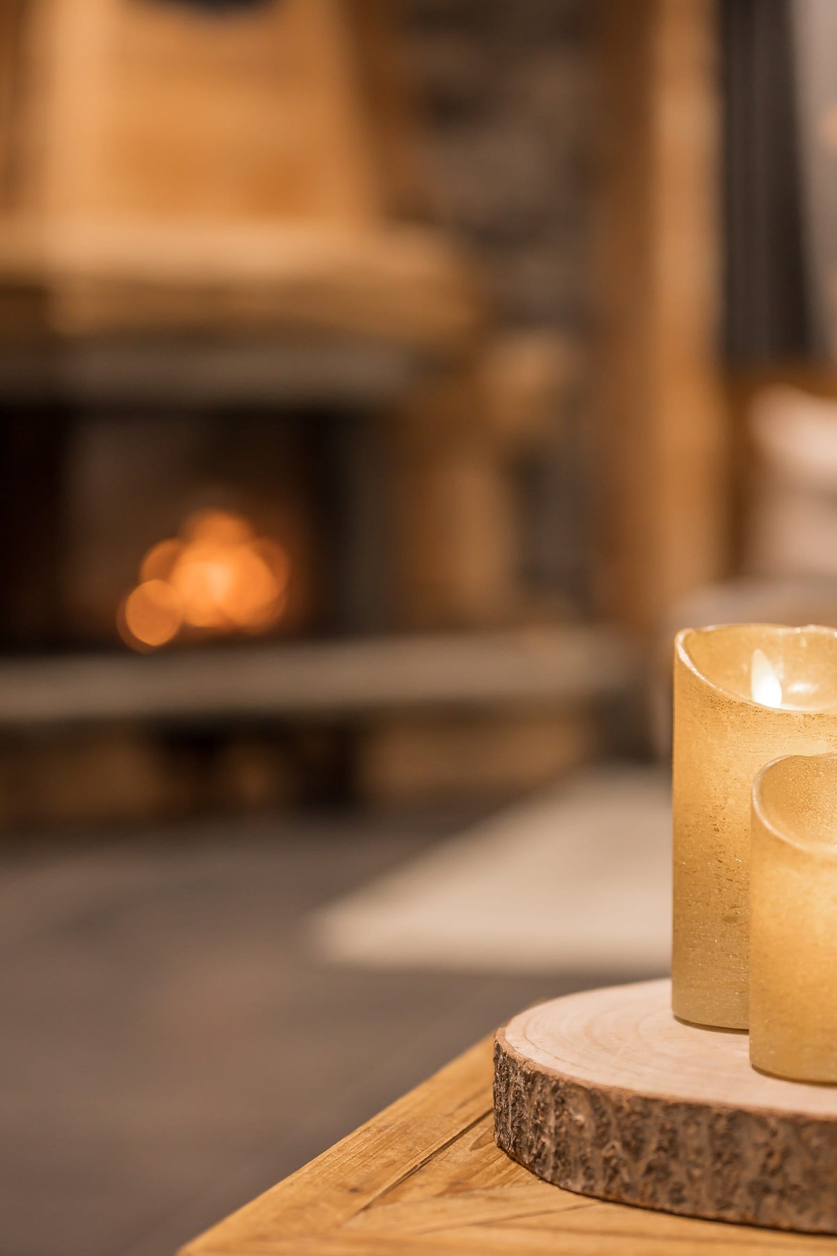 Three lit flameless LED pillar candles in warm cream and beige tones, arranged on a round wood slice coaster on a wooden table in a cozy, warmly lit cabin living room with a fireplace in the background.