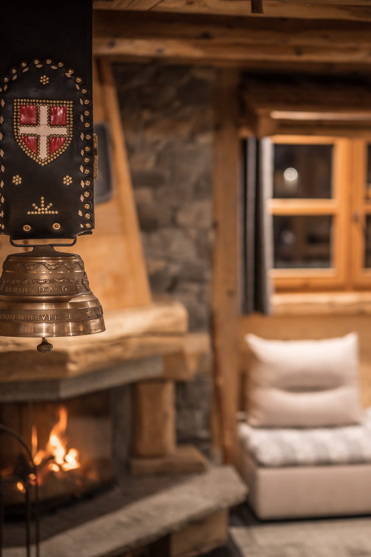 A rustic wooden chalet living room with a stone fireplace, a hanging brass bell ornament with a decorative crest, a low white sofa with cushions, and a wooden coffee table in the foreground.