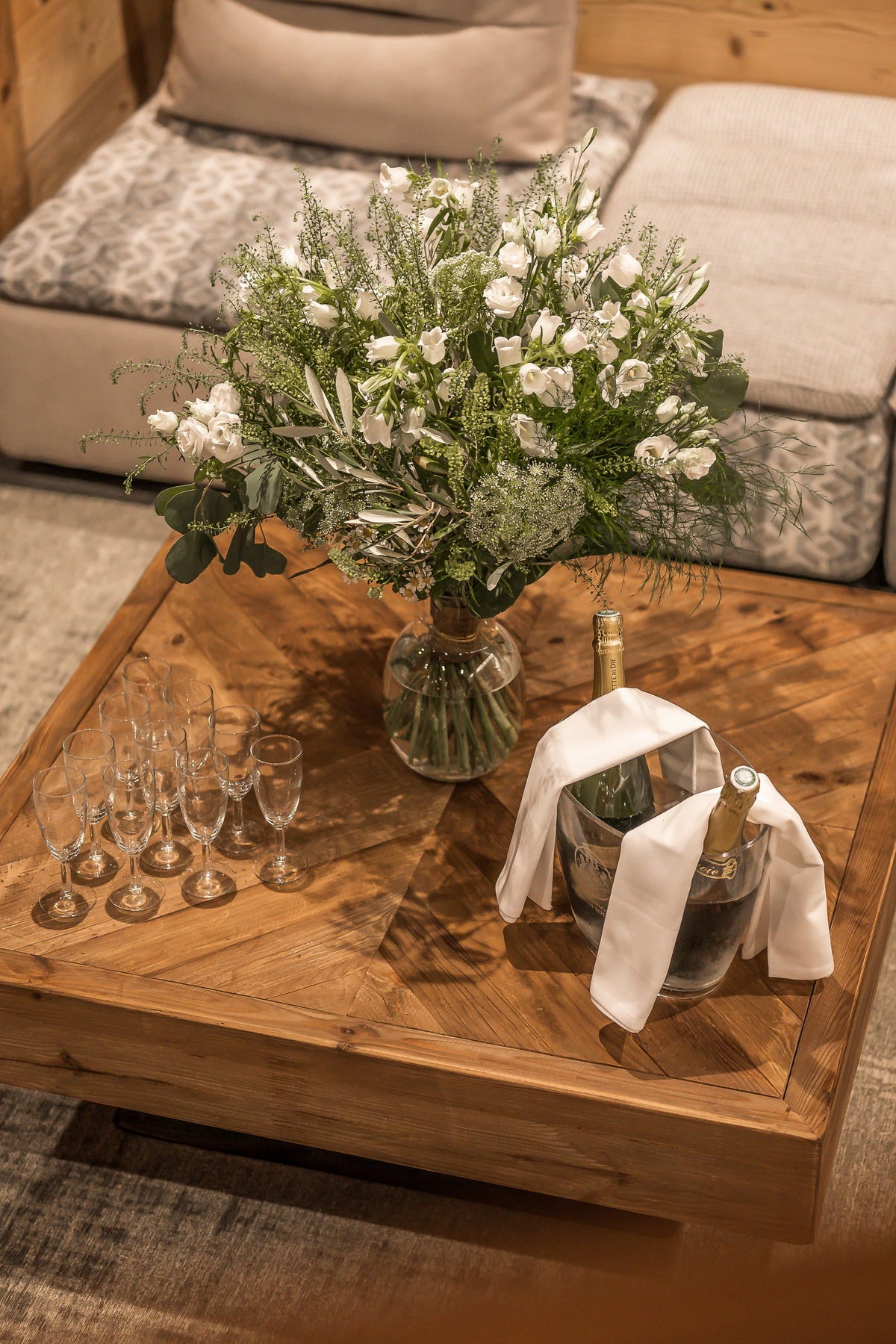 A rustic wooden coffee table in a cozy living room setting, styled with a large vase of white flowers, several wine glasses, and an ice bucket with a white cloth and gold bottle.