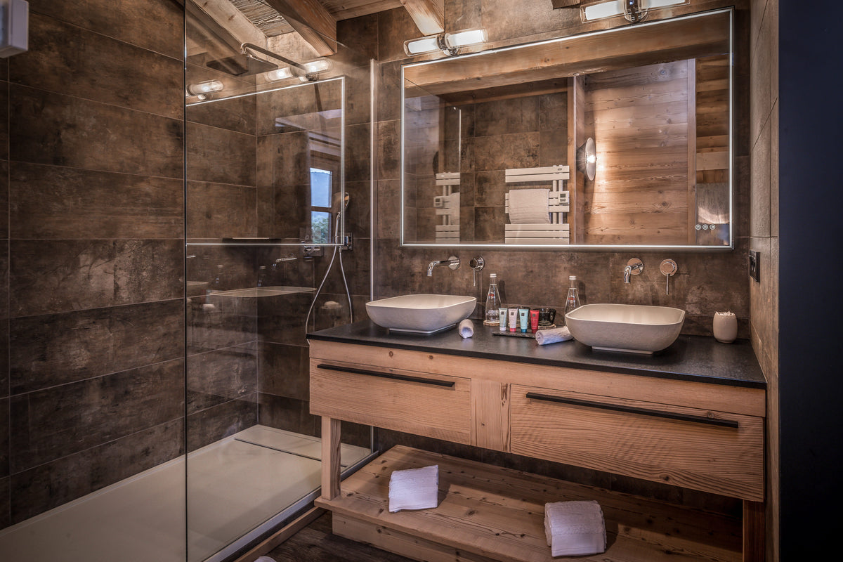 A luxury modern bathroom with a double vessel sink vanity, large illuminated mirror, dark stone countertop, rustic wood walls, and a glass walk-in shower enclosure.