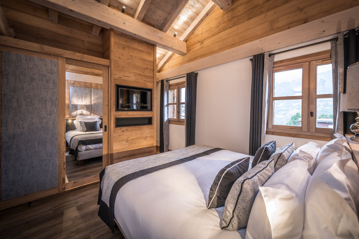 A modern mountain chalet bedroom featuring a large bed with white linens and gray patterned pillows, set in a rustic wooden room with a built-in TV, sliding door, and windows looking out to a scenic landscape.