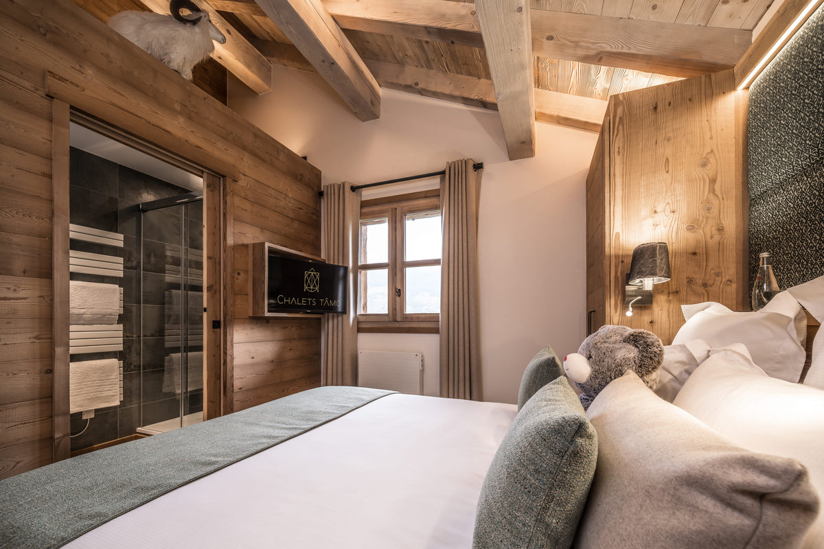 A luxury bedroom interior with a large bed in the foreground, rustic wood-paneled walls and ceiling, a window with curtains, and a wall-mounted TV. The room is warmly lit and styled for comfort.