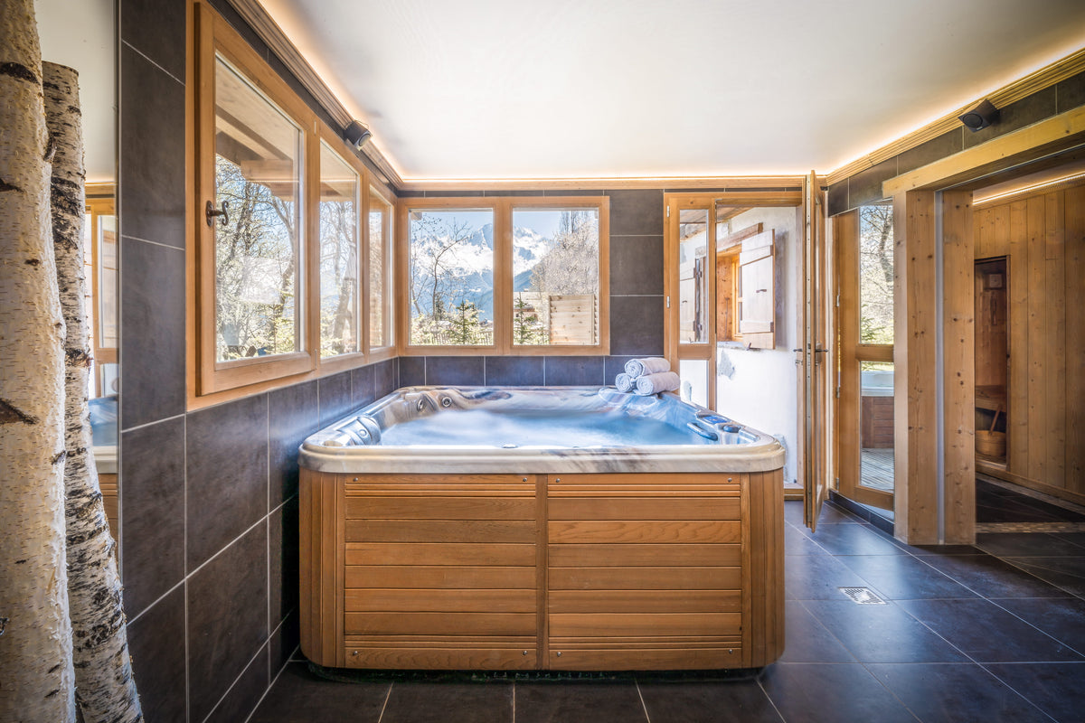 A luxury wooden hot tub with a marble-patterned rim, situated in a modern chalet bathroom with dark tiled walls and large windows overlooking a mountain view.