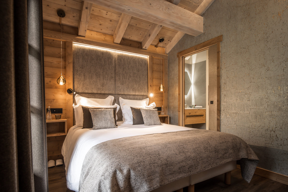 A luxury bedroom featuring a large bed dressed in white linens and textured gray pillows, set against a rustic wood-paneled wall with an integrated LED light strip. The room has a high vaulted ceiling with exposed wooden beams and a warm, inviting ambiance.