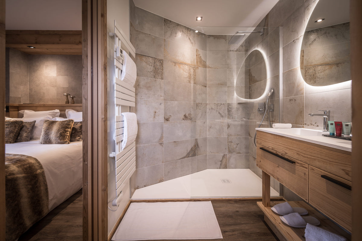 A luxury modern bathroom featuring a walk-in shower with stone tile walls, a wooden vanity with integrated sink and illuminated mirror, a white bath mat, and a glimpse into an adjoining bedroom through a wooden door.
