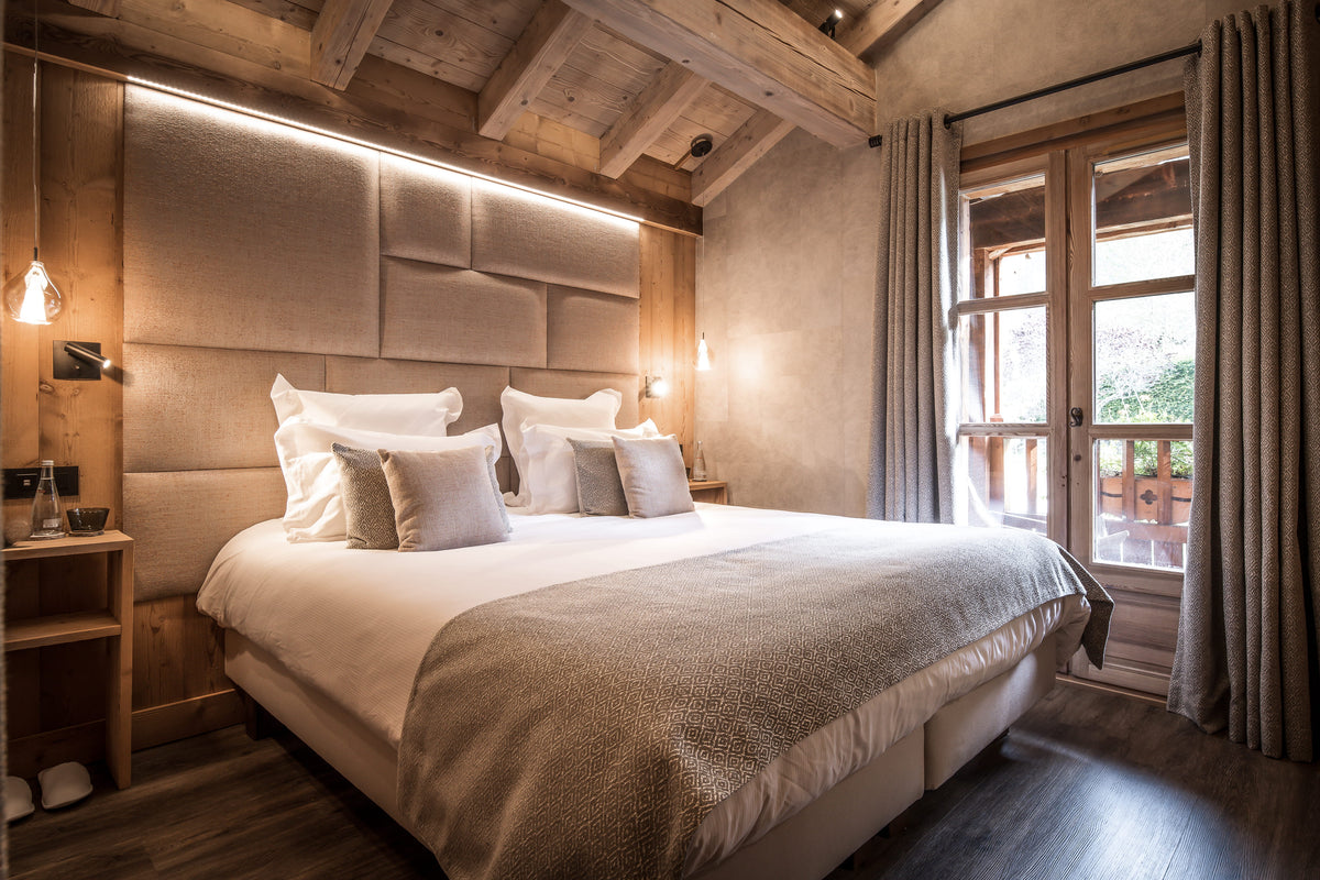 A luxury bedroom featuring a large bed dressed in white linens and a textured beige throw, set against a padded wood headboard with integrated lighting. The room has rustic wooden ceiling beams, a wooden nightstand, and a window with curtains looking out to a balcony.