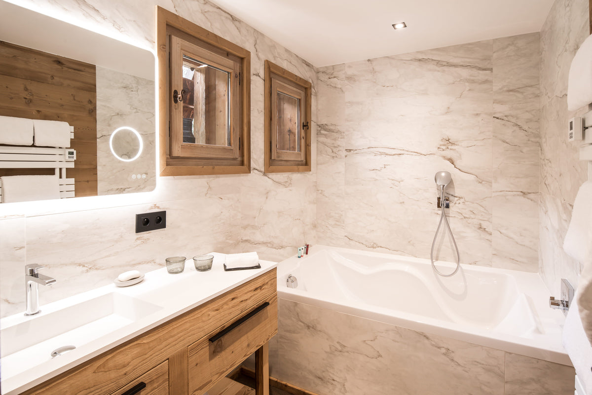 Luxury modern bathroom featuring a white marble bathtub, a double-sink vanity with light oak wood cabinets, a large illuminated mirror, and two matching wood-framed wall mirrors.