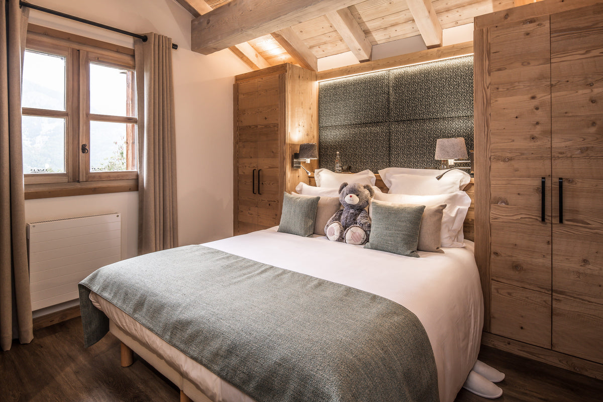 A cozy bedroom interior featuring a neatly made bed with white linens, gray-green throw and pillows, a plush toy, a dark textured headboard, wooden ceiling beams, a window with curtains, and a large wooden wardrobe.