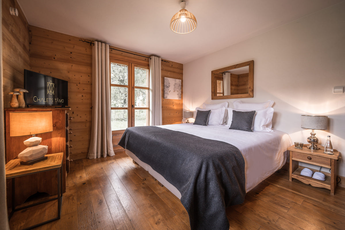 A cozy, rustic bedroom with a double bed dressed in white and dark gray bedding, set against wood-paneled walls with a window, bedside tables, and warm ambient lighting.