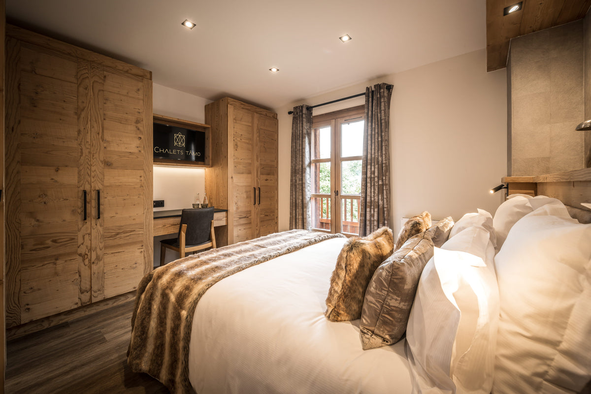 A rustic bedroom interior with a large bed dressed in white linens and patterned pillows, wooden wardrobes, a desk nook, and a window with curtains.