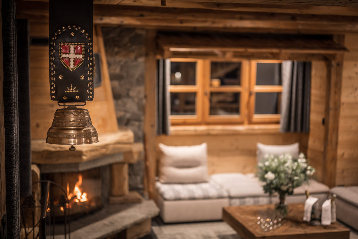 A rustic wooden chalet living room with a stone fireplace, a hanging brass bell ornament with a decorative crest, a low white sofa with cushions, and a wooden coffee table in the foreground.