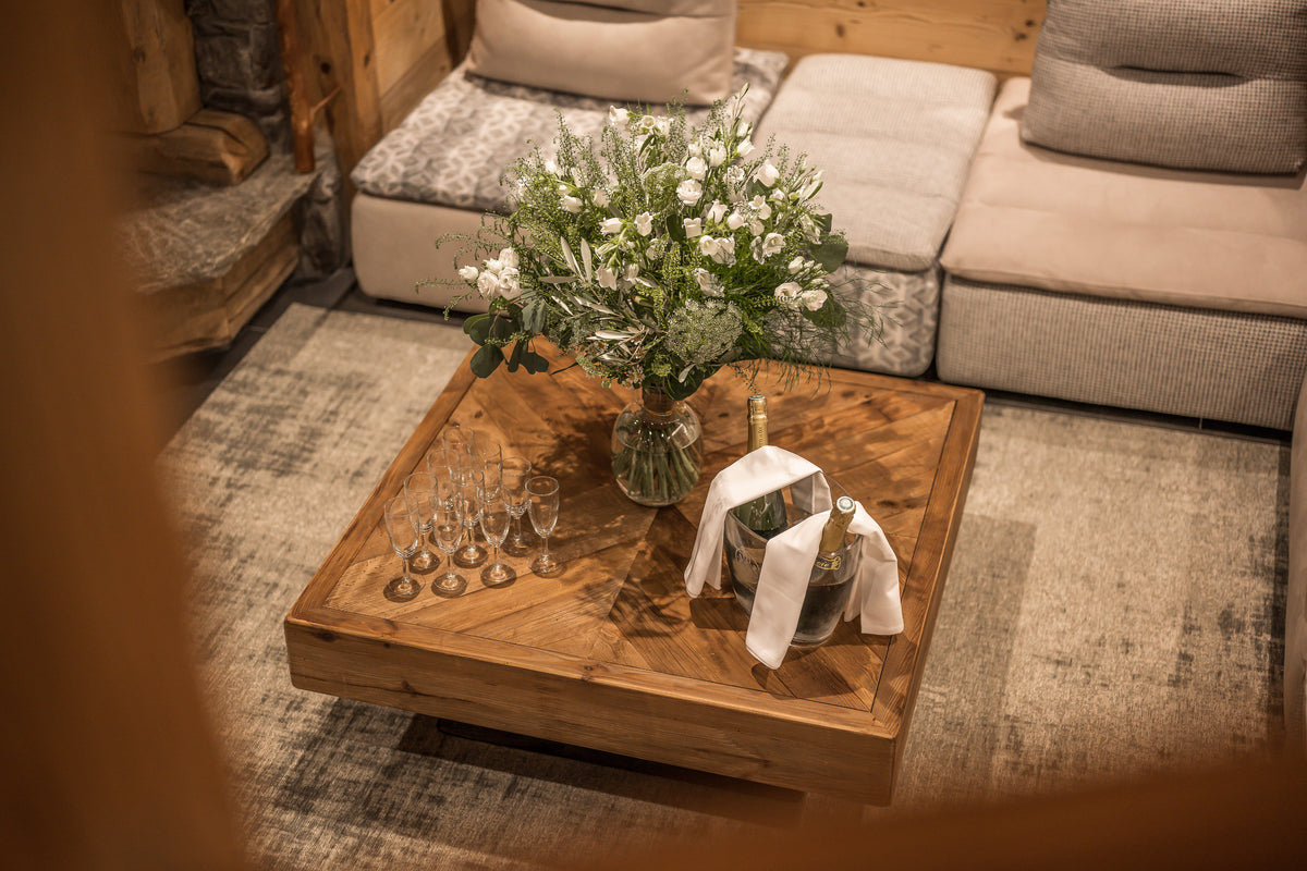 A rustic wooden coffee table in a cozy living room setting, styled with a large vase of white flowers, several wine glasses, and an ice bucket with a white cloth and gold bottle.