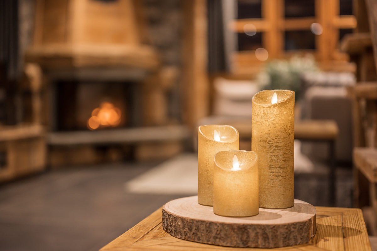 Three lit flameless LED pillar candles in warm cream and beige tones, arranged on a round wood slice coaster on a wooden table in a cozy, warmly lit cabin living room with a fireplace in the background.