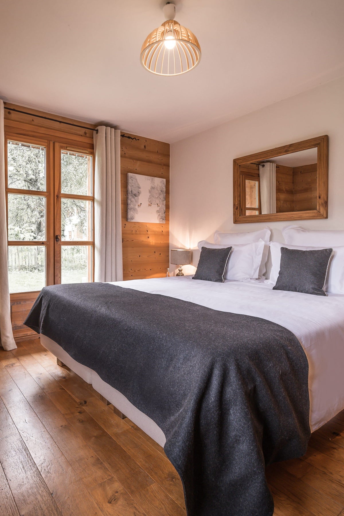A cozy, rustic bedroom with a double bed dressed in white and dark gray bedding, set against wood-paneled walls with a window, bedside tables, and warm ambient lighting.