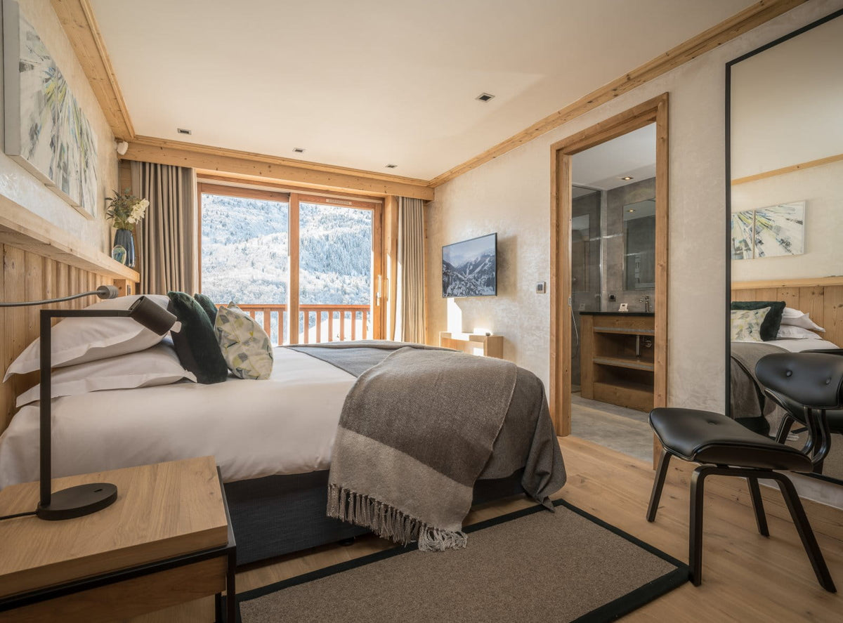 A modern mountain-view bedroom featuring a neatly made bed with white linens and a gray throw blanket, light wood walls and ceiling trim, a wall-mounted TV, a black chair, and a large mirror reflecting the room. A bathroom is visible through an open doorway in the background.