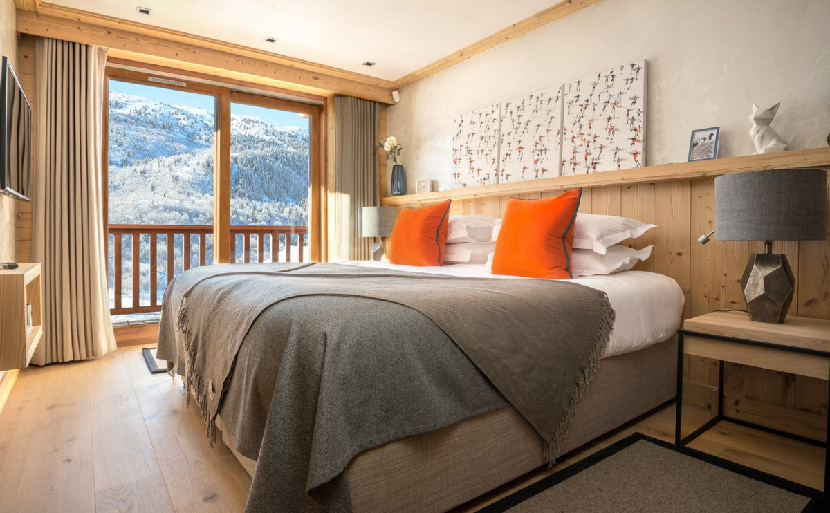 A modern mountain-view bedroom with a large bed dressed in gray and white bedding, bright orange accent pillows, light wood furniture, and floor-to-ceiling windows showing snowy alpine scenery.