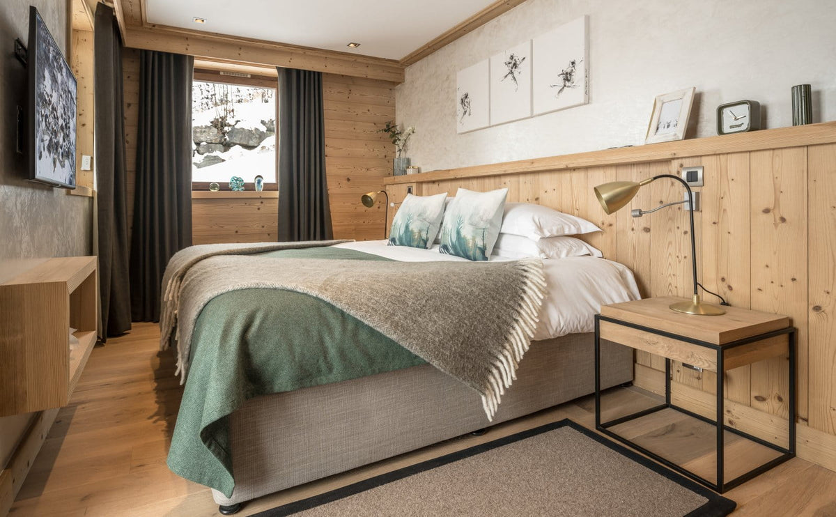 A modern bedroom interior with a neatly made bed, green and beige throw blanket, white pillows, light wood paneled walls, a bedside table with a brass lamp, and a window with dark curtains.