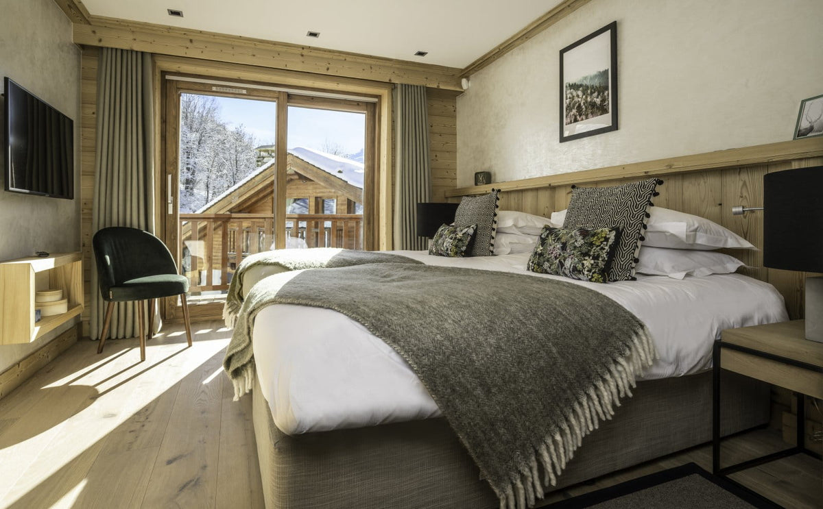 A cozy mountain chalet bedroom featuring a large bed with white linens and a gray knit throw blanket, set in a warm wood interior with a large window overlooking a snowy outdoor mountain scene.